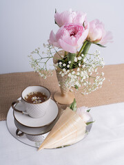 Artistic still life with a bouquet of pink peonies and a cup of tea. Pink peony flower in a horn. Flowers for design. Vertical photo.