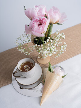 Artistic still life with a bouquet of pink peonies and a cup of tea. Pink peony flower in a horn. Flowers for design. Vertical photo.