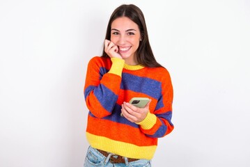Joyous Young caucasian woman wearing colorful knitted sweater poses with mobile phone device, types text message on modern smartphone, watches funny video during free time, enjoys good internet