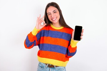 Young caucasian woman wearing colorful knitted sweater over white background holding in hands cell showing ok-sign