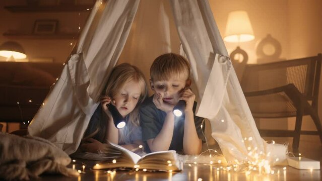 little kids read book with flashlights in handmade teepee from blanket in living room