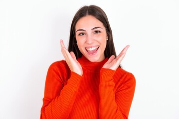 Surprised happy Young caucasian woman wearing red sweater over white background , glad to see big discounts on clothes, expresses shock, keeps hands near head, jaw dropped.