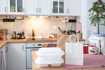 Fototapeta na wymiar Food delivery service containers on table white scandi festive kitchen in christmas decor. Eve New year, saving time, too lazy to cook, hot order, disposable plastic box in fairy light. mock up
