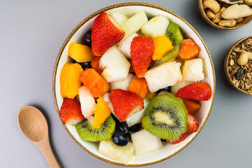 Fresh fruit salad in a bowl. Multicolored and tropical fruits. Pineapple, mango, grape, strawberry, papaya, melon, kiwi. Additional with chestnuts and granola. Top view. Selective focus