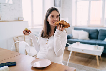 Happy expectant woman eating croissant in kitchen at home