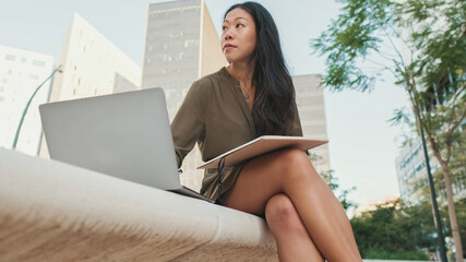 Young girl uses laptop pc makes notes in notebook from outside on modern buildings background
