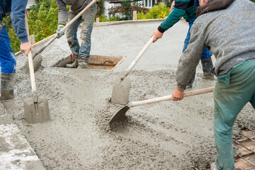 WORKERS ARE EVENING A LAYER OF CEMENT SOLUTION WITH SHOVELS AFTER POURING THE SOLUTION FROM THE CONTAINER