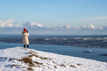 Woman with a red hat taking a picture with the phone of the views from the mountain. Cave Hill mountain with snow in winter. Belfast, Nothern Ireland
