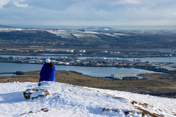 Woman listening to music and loking at the horizon on the snow. Person sitting and chilling on the mountain. Views over Belfast harbor from Cave Hill in winter. Northern Ireland.