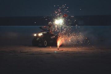 Athlete on a quad bike rides at night on a sandy beach with fireworks