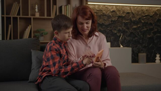 Mother learning her autistic son how to use smartphone sitting on the sofa at home. Boy with autism using mobile phone with mom's help.