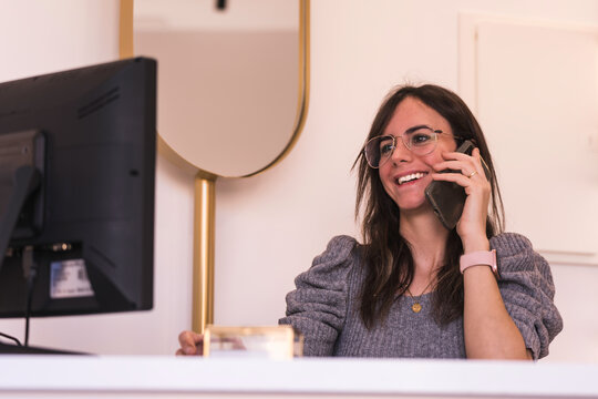 brunette receptionist with glasses smiles while using her computer and talking on her smartphone