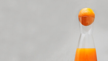 a narrow bottle with bright orange juice on a light gray background, there is a whole orange in the neck of the bottle