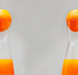 two oranges are half visible in the neck of narrow glass bottles with bright orange juice on a light gray background along the edges of the image