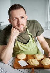 Portrait of caucasian baker man in green apron uniform smiling looking at camera. Portrait of handsome baker at home sitting at the table with dough. High quality vertical photo