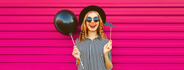 Portrait of funny cheerful young woman having fun holding black balloon, showing mustache on stick...