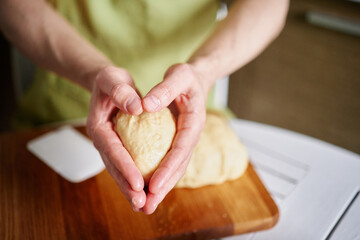 Working at home kitchen concept, homemade baking. Man chef baker in green apron holding dough ball in hands folded in heart shape. High quality photo
