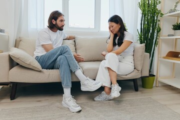 A sad Asian woman talks to a man in tears at home sitting on the couch. Young couple of different nationalities and conflict of interest in a couple