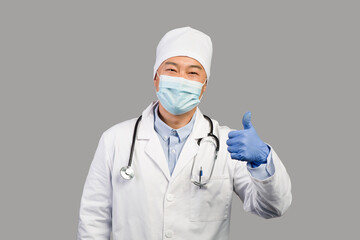 Smiling middle aged chinese male therapist in white coat, protective mask with stethoscope showing thumb up