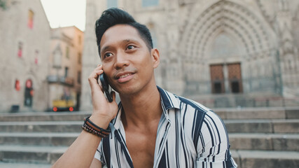 Fototapeta premium Young man tourist sitting on the steps talking on mobile phone with the sights in the background