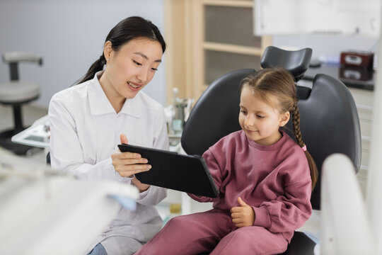 Medicine, pediatric dentistry and oral care concept. Female smiling asian dentist showing tablet pc computer to happy kid patient, caucasian preschool girl at modern dental clinic.