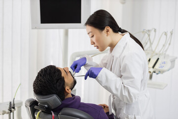 Beautiful smile with white teeth. Asian dentist treating teeth of a young bearded man using tooth...