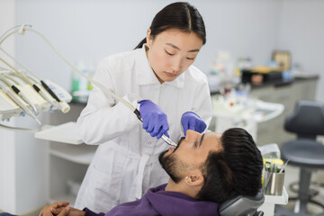 Happy patient and dentist concept. Young beautiful asian female stomatologist treating teeth of a handsome bearded young guy, using tooth drill. Oral health and hygiene.