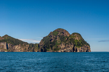 Fototapeta premium Resurrection Bay, Alaska, USA - July 22, 2011: double boulder-like end of land at ocean under blue sky. Green foliage tries to cover the brown rocks but cliffs stop it