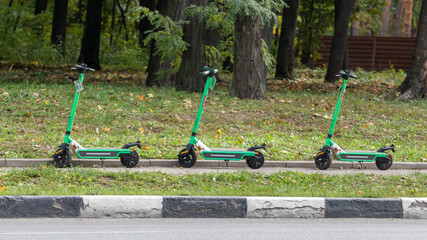 Green Electric Scooters rental standing in city street in greenery. Public e-scooter, popular modern transportation