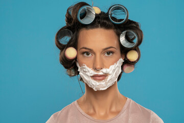 attractive woman with hair curlers rollers posing with shaving foam on her face on a blue background