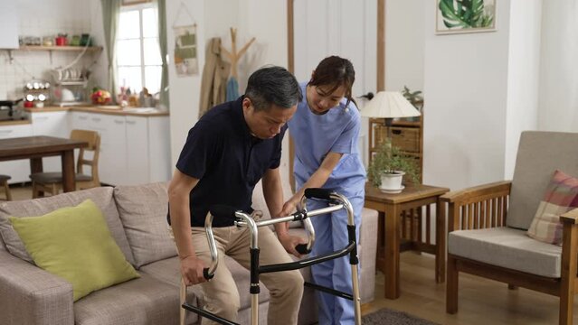 slow motion of asian older male stroke patient practicing using a walker with the assistance of his personal care attendant in the living room at home