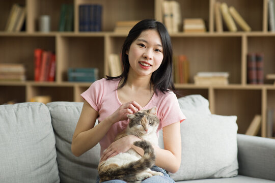 Portrait of happy asian lady enjoying spending time with her cat at home, sitting on sofa and smiling at camera