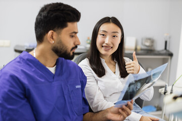 Attractive young asian woman, sitting on dentist chair and looking at x-ray picture of scan image of teeth together with her confident professional male dentist at clinic showing thum up. - obrazy, fototapety, plakaty