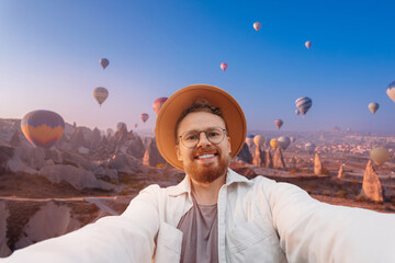 Young hiker man in hat taking selfie portrait on top mountain background hot air balloons in Cappadocia Turkey travel. Happy guy smiling at camera