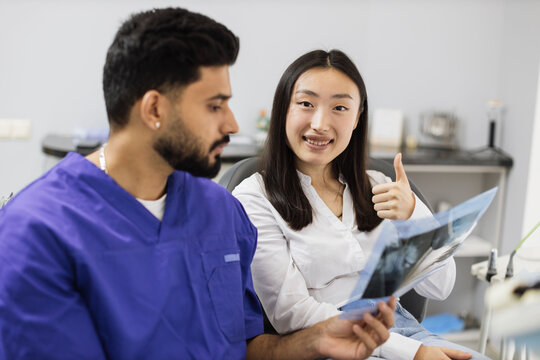 Attractive young asian woman, sitting on dentist chair and looking at x-ray picture of scan image of teeth together with her confident professional male dentist at clinic showing thum up.