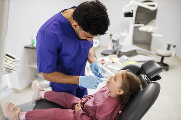 Beautiful smile with white teeth. Confident bearded dentist examines the oral cavity of a young beautiful girl through a magnifying glass in the dental office.