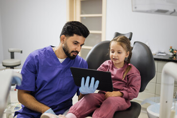 Medicine, pediatric dentistry and oral care concept. Confident male smiling professional dentist showing tablet pc computer to happy kid patient, caucasian preschool girl at modern dental clinic. - Powered by Adobe