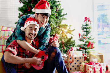 Obraz na płótnie Canvas Adorable young LGBT couple sharing special moment together on Christmas holiday, Asian gay male lover singing song while playing ukulele in living room Merry Christmas happy winter holiday celebration