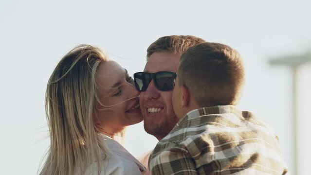 Portrait close-up of a happy smiling family father, mother and son hugging kissing dad in sunglasses on the cheeks in nature, outdoors