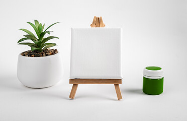 Blank clean white canvas on wood mini easel, mockup on wood tripod, green paint gouache jar and...