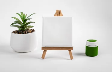 Fotobehang Blank clean white canvas on wood mini easel, mockup on wood tripod, green paint gouache jar and potted plant composition © valiantsin