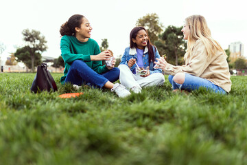 Three young female students having lunch sitting on grass at campus college