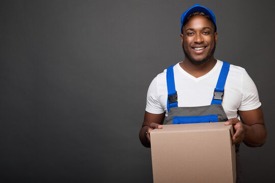 Cheerful African deliveryman easily holds a heavy cardboard box in his hands. Black man loader in overalls with a white T-shirt and a blue cap holds a cardboard box with a parcel in front of him.