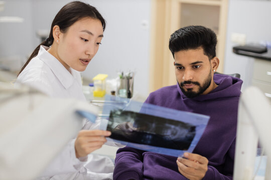 Attractive young bearded man, sitting on dentist chair and looking at x-ray picture of scan image of teeth together with his cheerful female asian dentist at clinic.