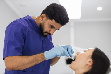 Fototapeta premium Side view of bearded male doctor in white uniform and gloves, making examination or curing caries and toothache for his female patient, attractive young asian woman sitting in dental chair.