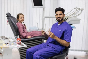 Children's teeth treatment. Smiling little girl sitting in the dentist's chair in light modern dental clinic to excited confident man dentist and looking at camera.