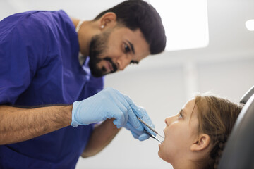 Fototapeta premium Side low angle view of smiling male professional doctor in blue uniform and gloves, making examination or curing caries and toothache for his little patient, cute kid girl, sitting in dental chair.