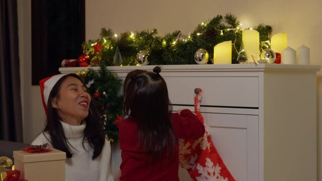 Asian family merry xmas and happy new year holiday. Mom and daughter enjoy beautiful time put present christmas put gift box decoration on red santa stocking hanging in living room night at home.