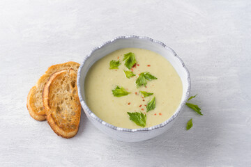 Cream soup of stalked celery with cream, garnished with celery leaves, pink pepper and served with crispy toast on a light gray background