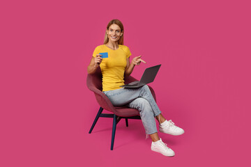 Fototapeta na wymiar Happy adult caucasian lady blonde in casual sits on armchair with credit card points finger at laptop